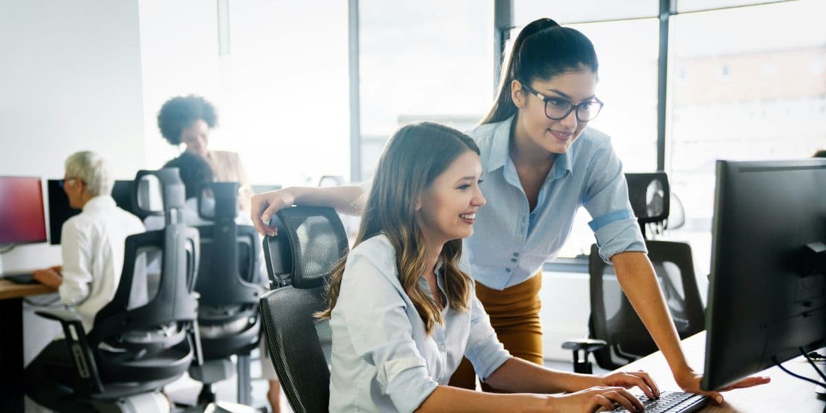 Zwei Frauen arbeiten gemeinsam am Computer, Kolleginnen und Kollegen im Hintergrund.
